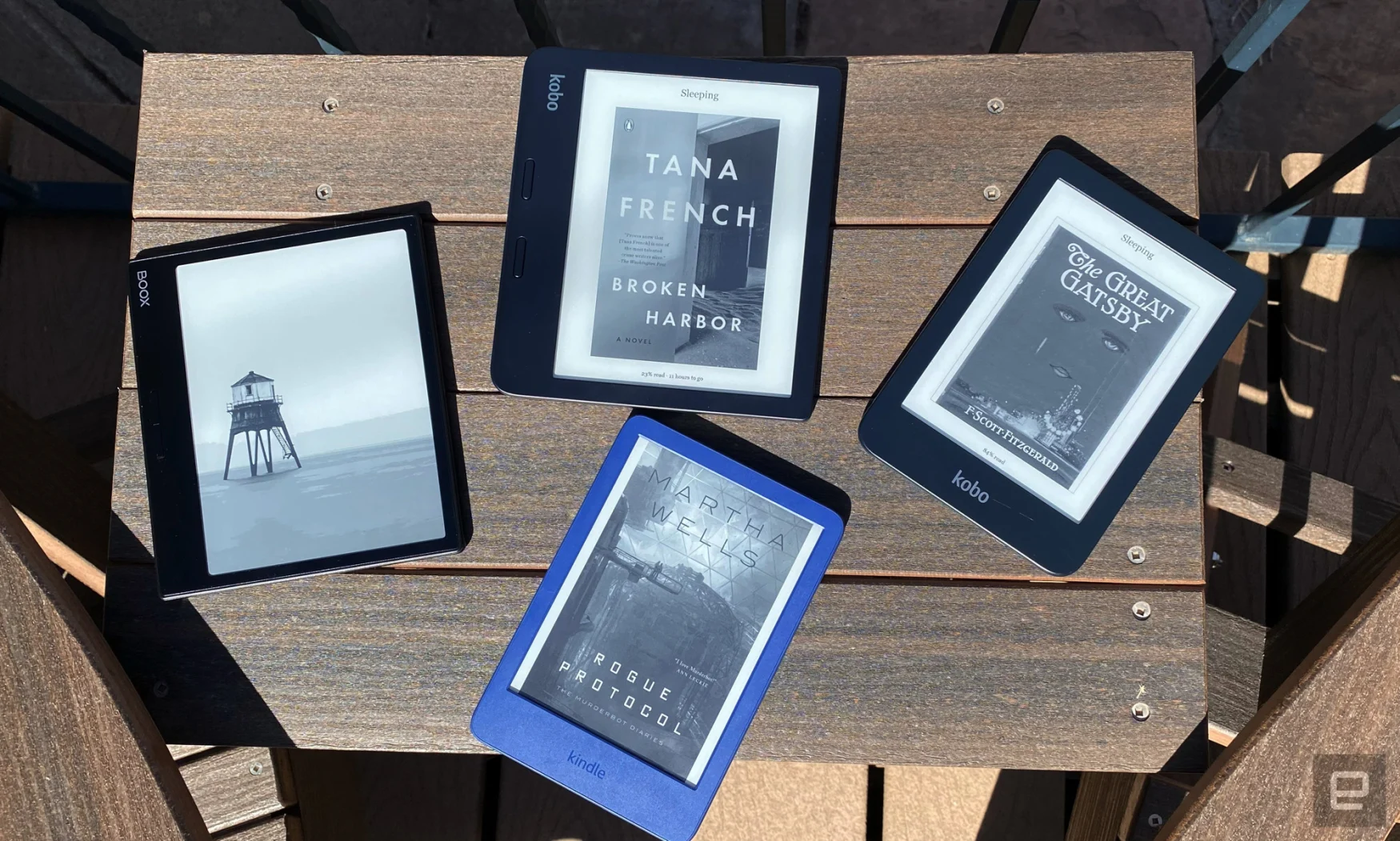 Four ereaders are arranged on a dark brown wooden table outside. There are Boox, Kindle and Kobo devices showing the covers of different novels from Tana French, F. Scott Fitzgerald and Martha Wells.  